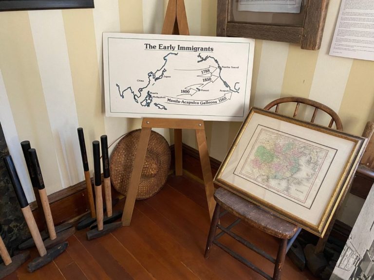 Photo of front display room showing the corner of the room to the left of the bay window with a map showing the movement of the early immigrants, tools, hat and map of China