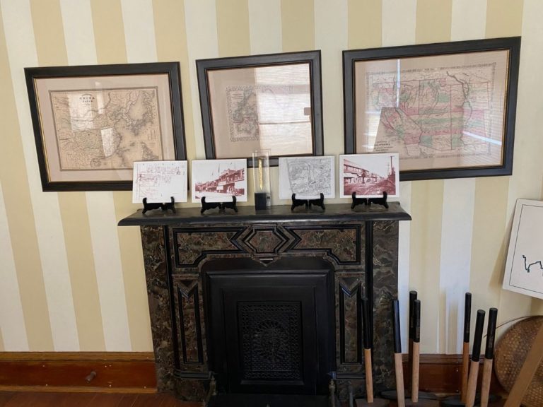 Photo of front display room showing the wall opposite the bay windows with framed maps on the wall, pictures of maps and buildings and our Golden Spike award on top of the fireplace