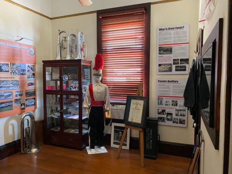 Photo of the Drum & Bugle Display room with an information poster on the left wall, display case, bugles, a mannequin dressed in uniform, more information posters on the back wall